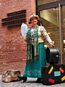Béatrice Forêt (Cie L’Arbre à Plumes) en costume d’époque devant l’entrée du Musée des Arts Précieux Paul-Dupuy, tenant un éventail blanc et entourée de valises anciennes.