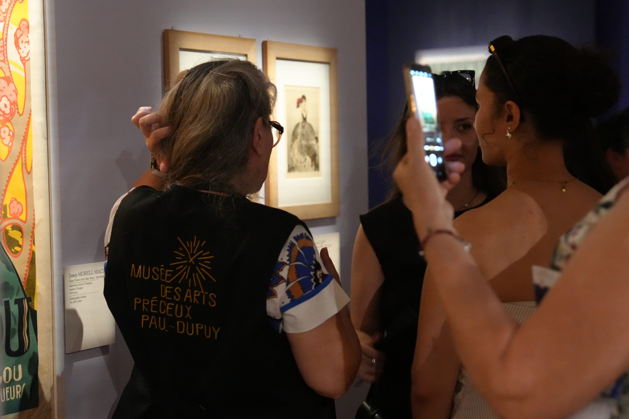 Visiteurs dans une salle de musée observant des œuvres encadrées accrochées au mur, tandis qu’une personne photographie la scène avec un téléphone portable.